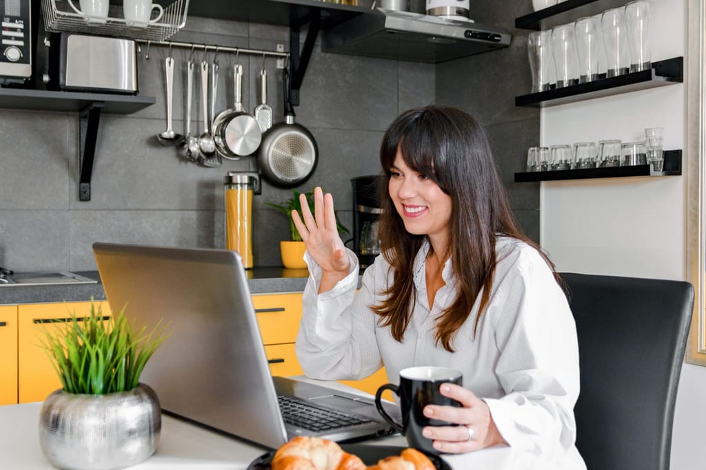 Photo of a woman in her modern kitchen talking on a video conference. She is holding a cup of coffee and seems very happy.